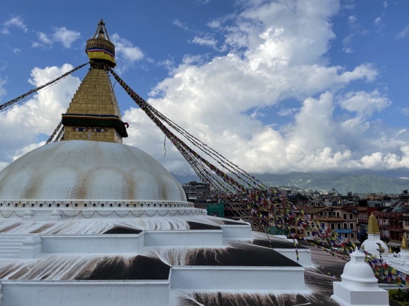Large white dome with golden spire on top, painted with a set of giant eyes and festooned with Tibetan prayer flags. Roogtops in the distance